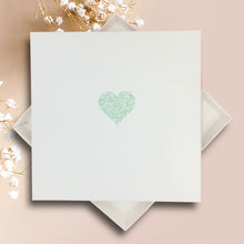 Load image into Gallery viewer, A white square card with a green glitter heart design is placed on a pink background.