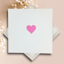 Load image into Gallery viewer, A white square card with a pink glitter heart symbol in the center, placed on a beige background with white flowers.