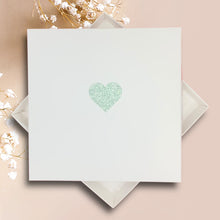 Load image into Gallery viewer, A white square card with a green glitter heart design is placed on a pink background.
